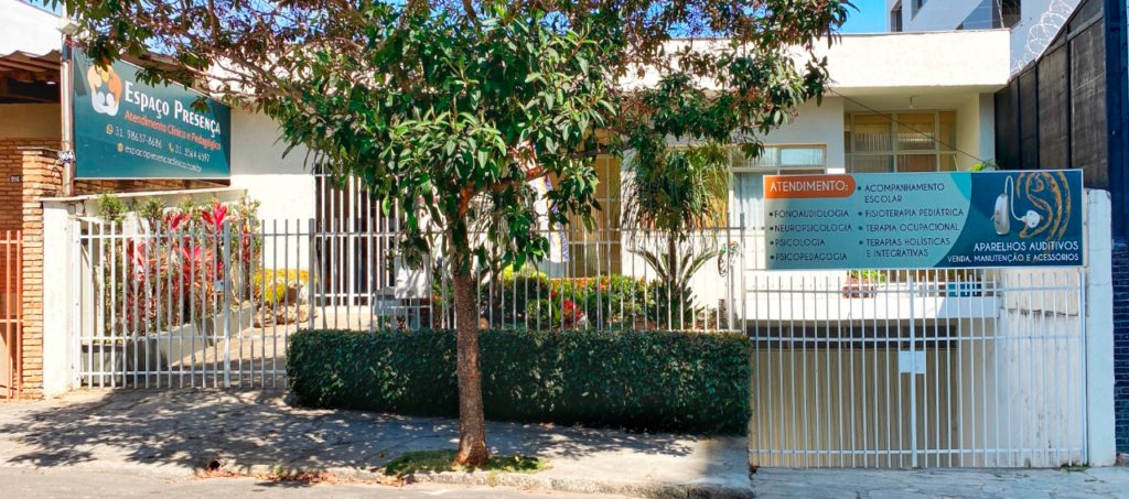 Fachada da clínica, com portão branco e árvore na calçada